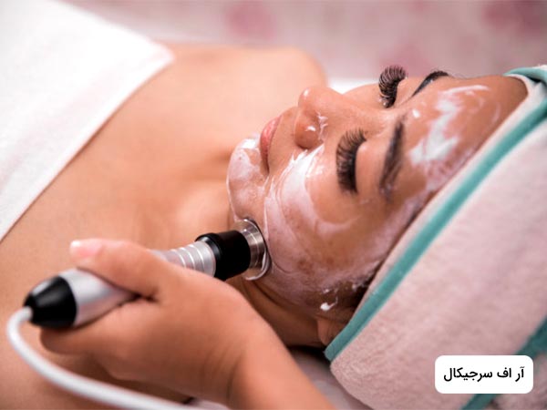 خانمي روي تخت دراز کشيده است. خانم روي صورت خود را کرم مخصوص انجام عمل راديو فرکانسي بع روش جراحي زده است. پزشک در حال درمان پوست صورت خانم با روش آر اف سرجيکال مي باشد.